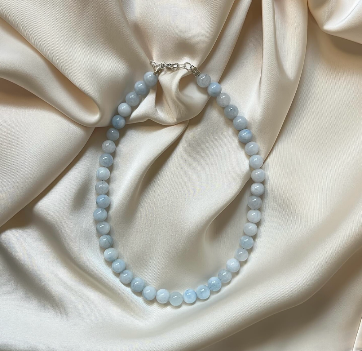 שרשרת ANGEL NECKLACE חרוזי אקוומרין תכלת מונחת על בד משי בצבע קרם, עם סוגר כסוף בולט.