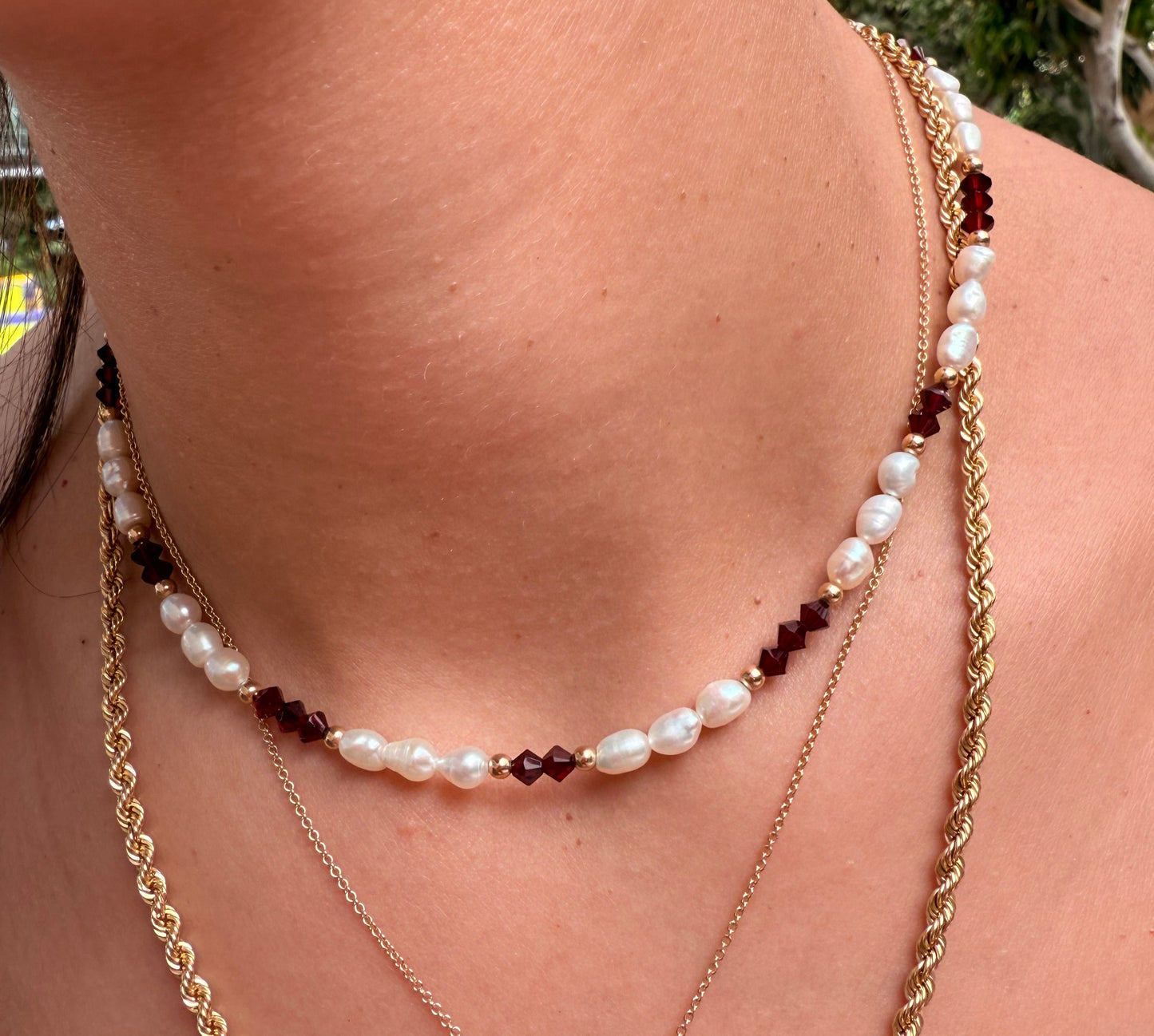 שרשרת ALAMOR BORDEAUX GOLD NECKLACE עם פנינים וחרוזי בורדו על צוואר דוגמנית, בשילוב שרשראות זהב בשכבות.