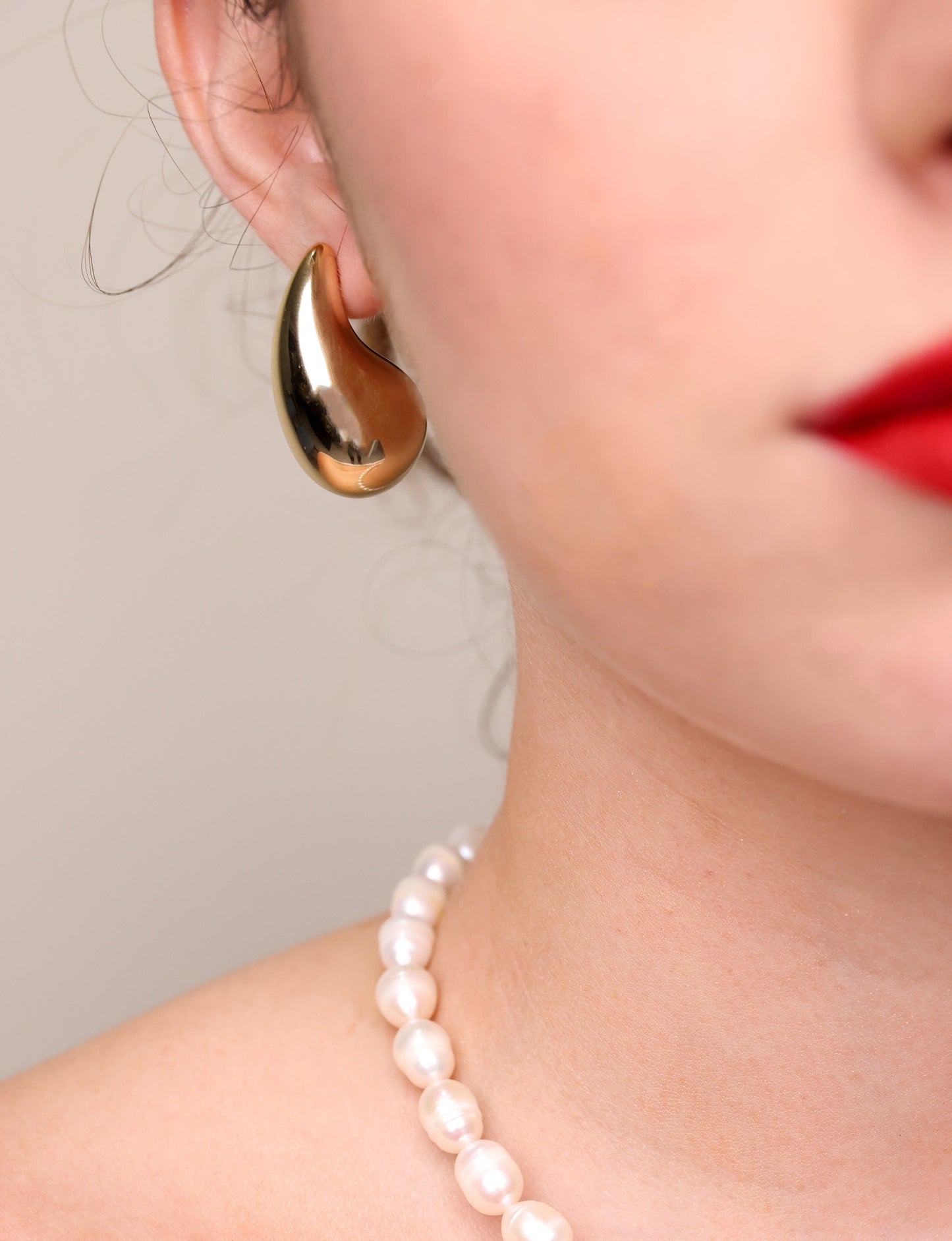 עגילי LOLA L זהב מעוצבים כטיפה מבריקה על אוזן דוגמנית, תקריב עם שפתון אדום ושרשרת פנינים.