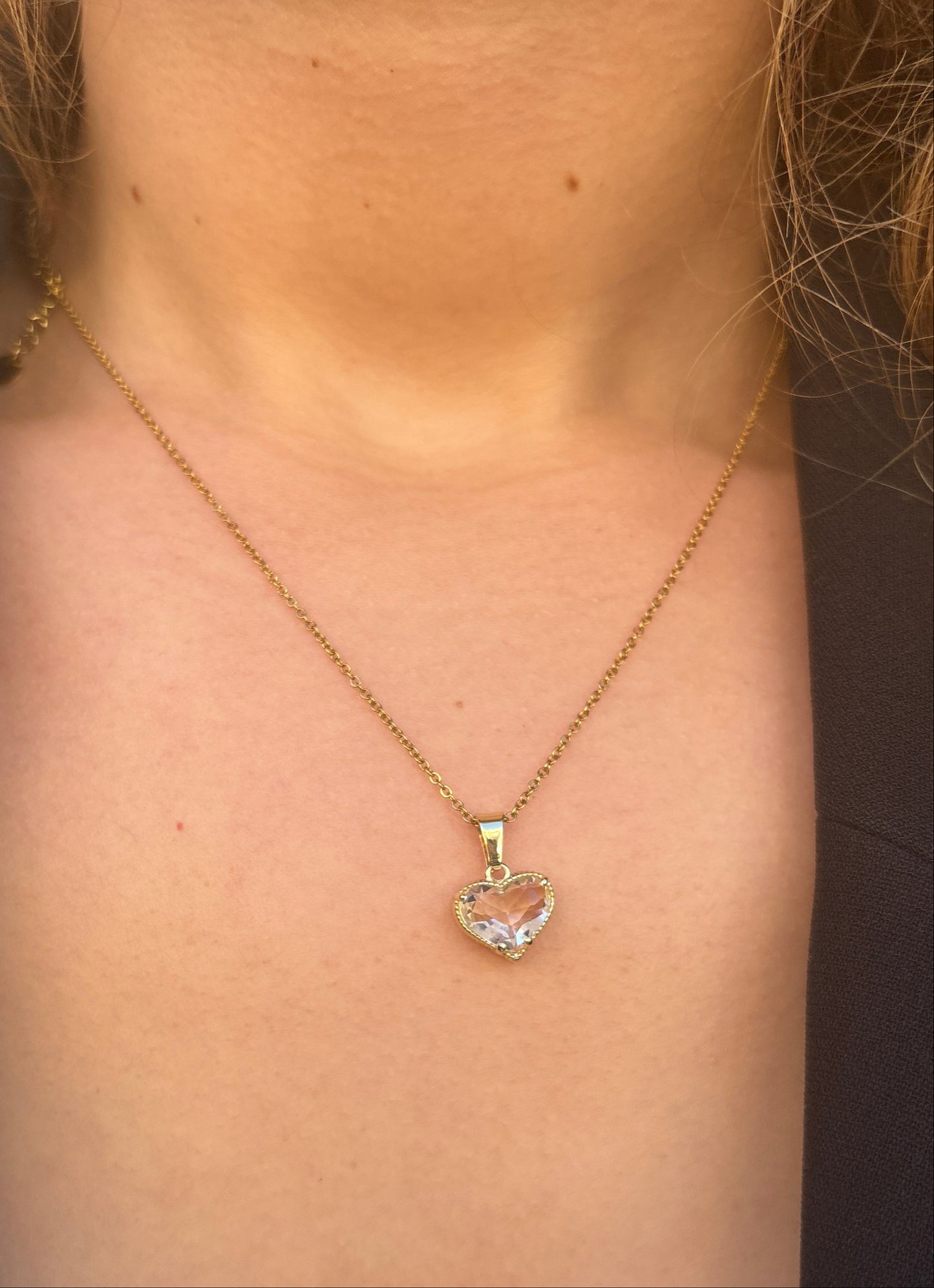 שרשרת LALYA NECKLACE זהב עדינה על צוואר דוגמנית, תליון לב קריסטל שקוף. תקריב על המחשוף עם שיער וז'קט כהה.