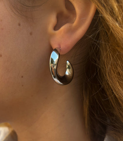 עגילי SHANI כסופים מבריקים, צורת קשת עבה על אוזן דוגמנית, תקריב עם השתקפות סביבה בעגיל.