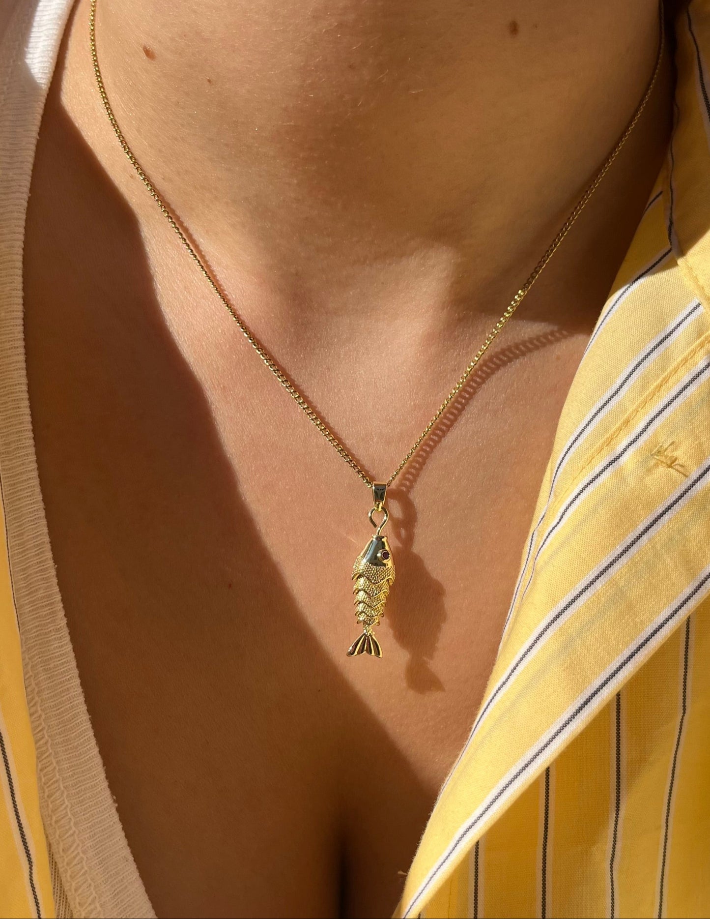 שרשרת POISSON זהב, תליון דג מפורט עם עין אדומה, על דוגמנית בחולצה צהובה מפוספסת.
