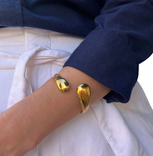 צמיד ETERNA BRACELET זהב מבריק, עם קצוות מלוטשים דמויי טיפה, ענוד על זרוע דוגמנית בלבוש כחול-לבן.