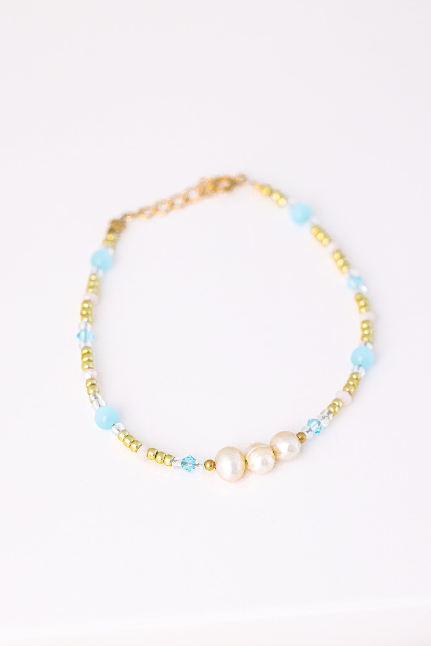 צמיד KARIN BRACLET מונח על רקע לבן, מעוטר 3 פנינים, חרוזי זהב, תכלת ושקופים, מבט עילי.