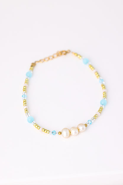 צמיד KARIN BRACLET מונח על רקע לבן, מעוטר 3 פנינים, חרוזי זהב, תכלת ושקופים, מבט עילי.