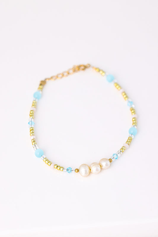 צמיד KARIN BRACLET מונח על רקע לבן, מעוטר 3 פנינים, חרוזי זהב, תכלת ושקופים, מבט עילי.