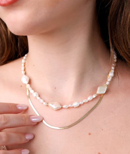 שרשרת SHAI NECKLACE בשכבות על צוואר דוגמנית, תקריב של פנינים לא אחידות ושרשרת זהב שטוחה, יד עדינה נוגעת.