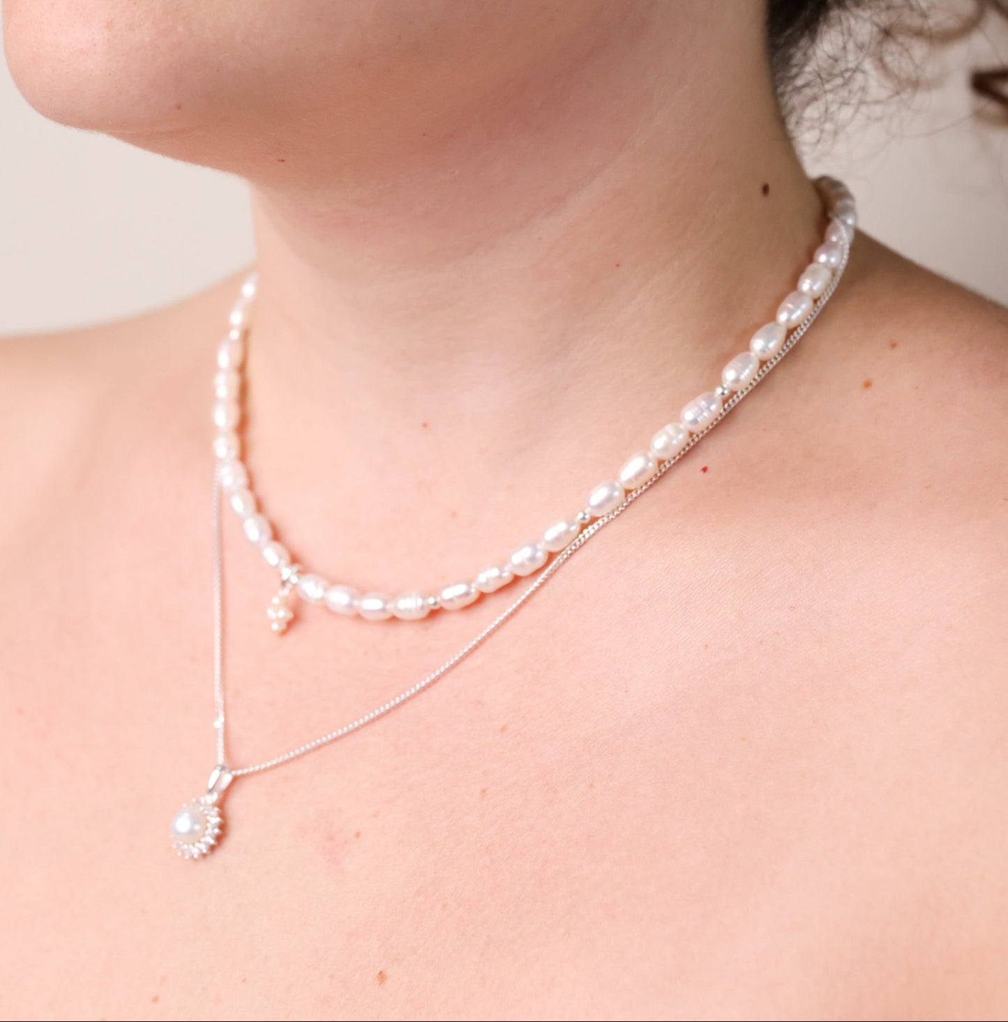 שרשרת NOA NECKLACE SILVER על צוואר דוגמנית, מראה שכבות: ה-NOA כתחתונה עם תליון פנינה וזרקונים, ועליונה שרשרת פנינים טבעיות.