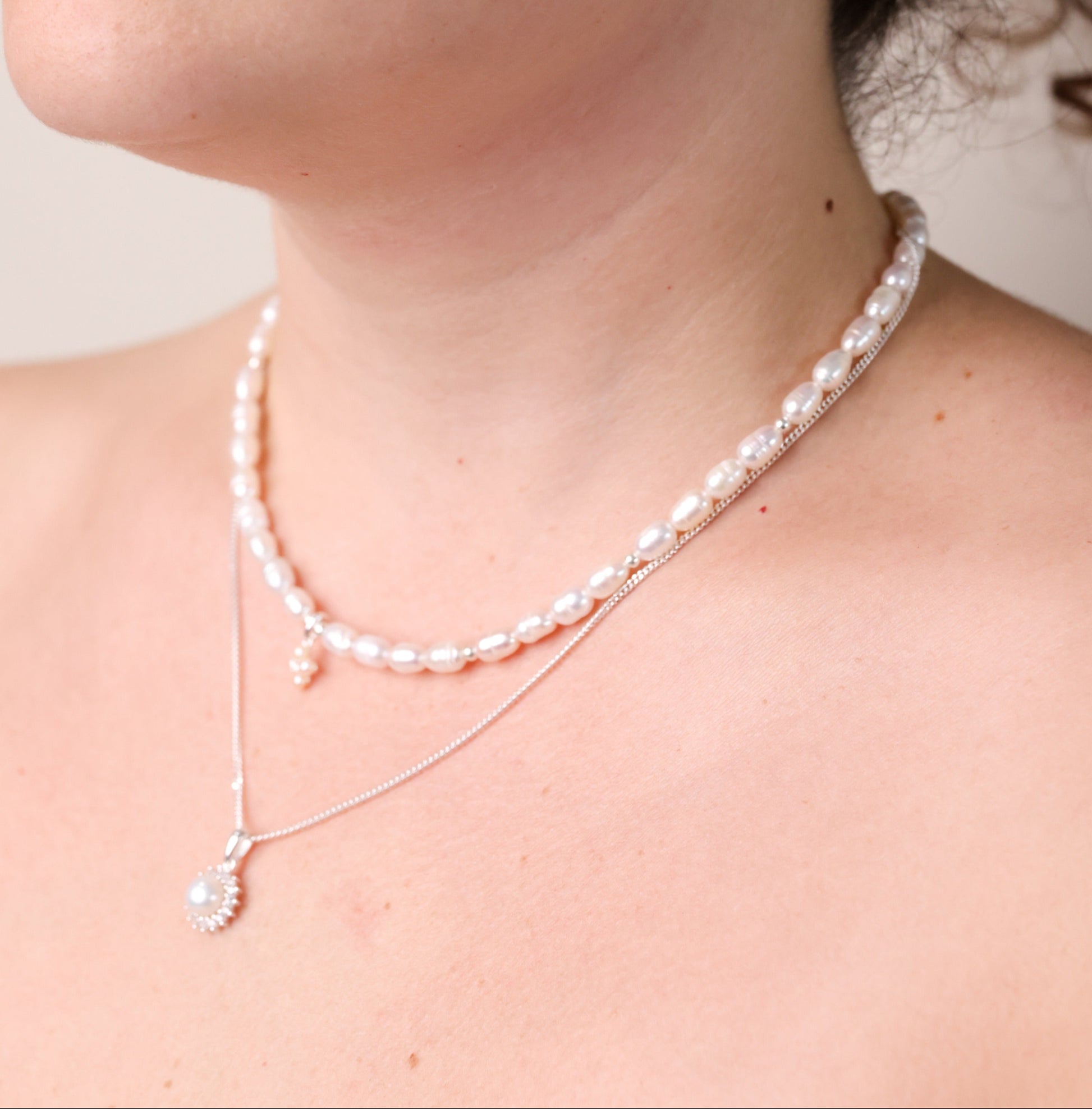 שרשרת NOA NECKLACE SILVER על צוואר דוגמנית, מראה שכבות: ה-NOA כתחתונה עם תליון פנינה וזרקונים, ועליונה שרשרת פנינים טבעיות.