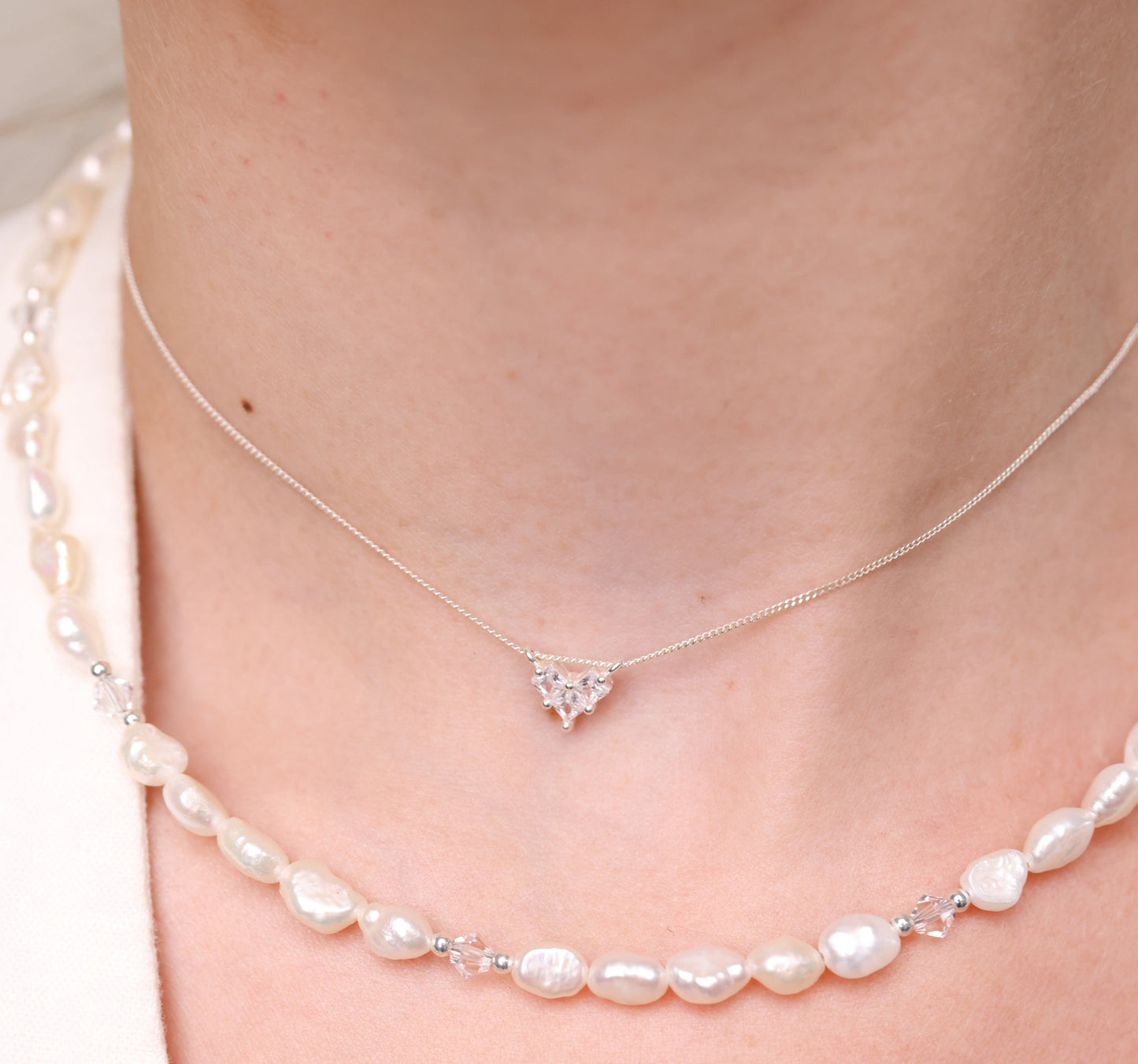 שרשרת ADELE NECKLACE כסף עדינה עם תליון לב משובץ, על צוואר דוגמנית לצד שרשרת פנינים במראה שכבות.