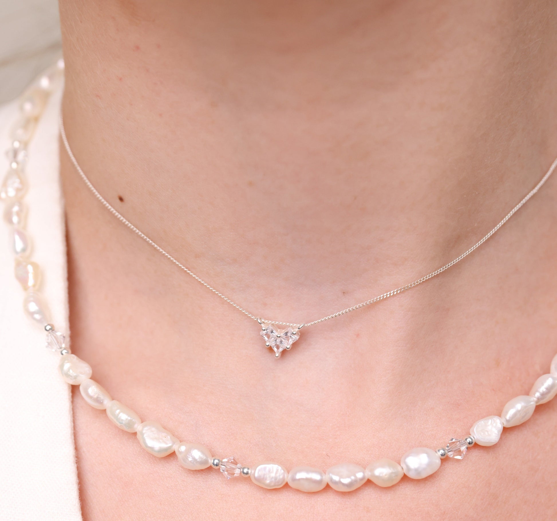 שרשרת ADELE NECKLACE כסף עדינה עם תליון לב משובץ, על צוואר דוגמנית לצד שרשרת פנינים במראה שכבות.