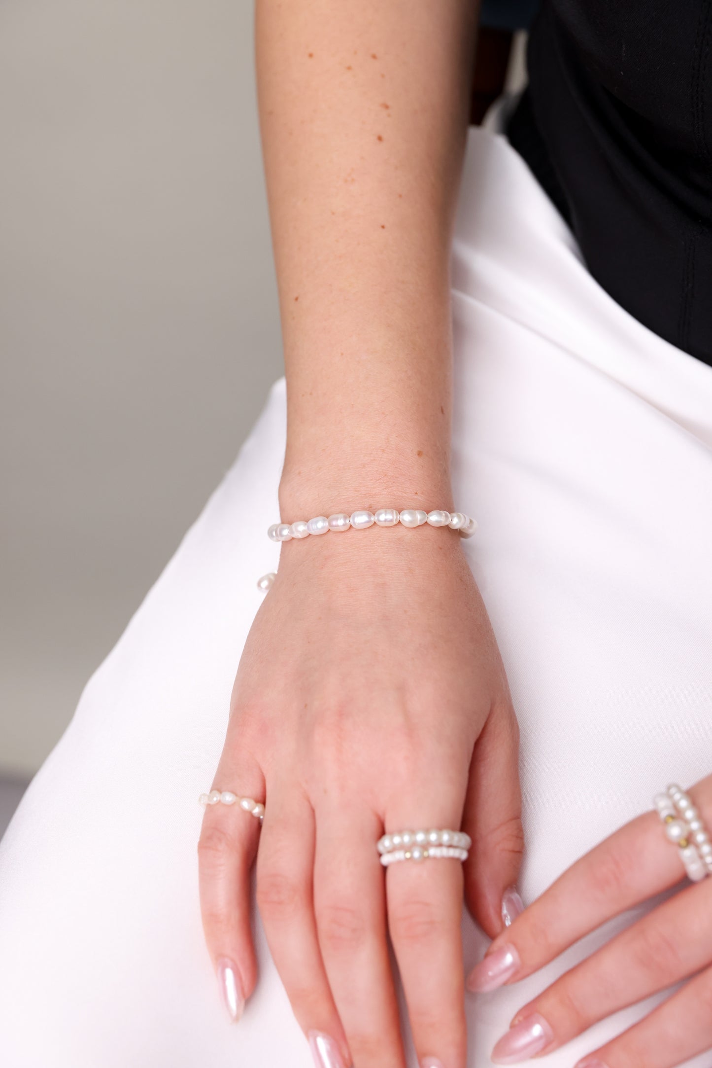 צמיד ANNA BRACELET פנינים על פרק יד דוגמנית, יחד עם טבעות תואמות על רקע בד סאטן לבן.