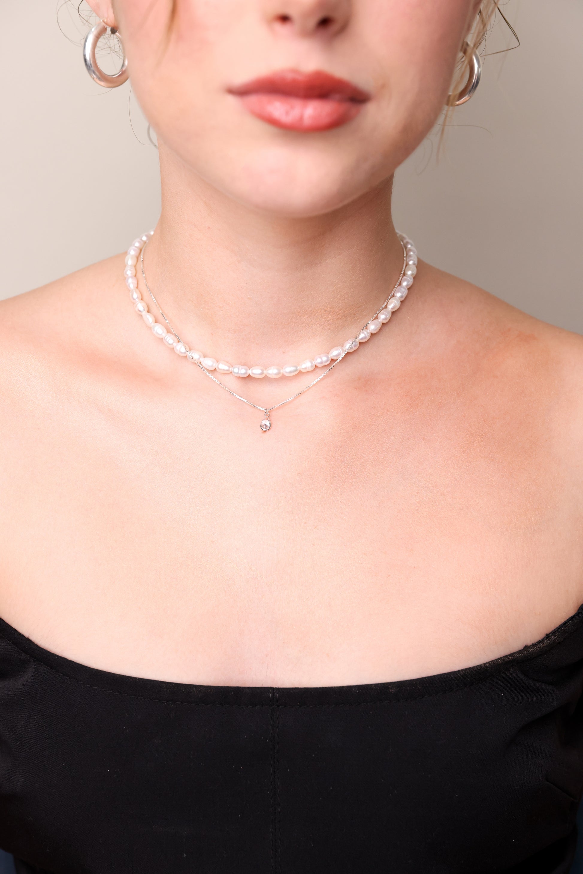 צוואר דוגמנית ותחילת מחשוף, עונדת שרשרת פנינים DARIA, משולבת בשרשרת דקה עם תליון קריסטל, תקריב.