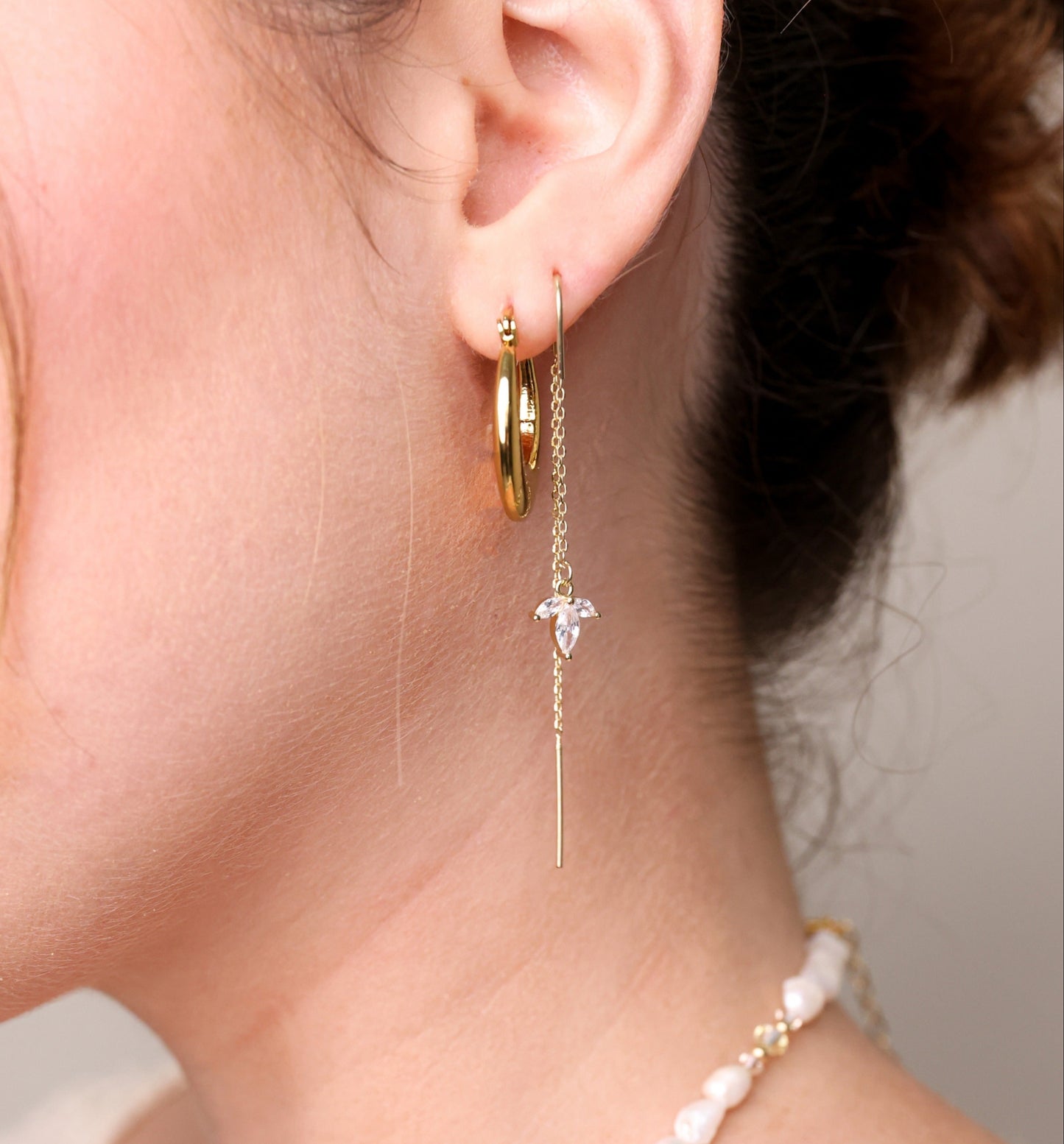 עגילי SARAI זהב על אוזן דוגמנית, תקריב מהצד: עגיל ארוך עם אבן שקופה ועמוד תלוי, ולצידו חישוק זהב.