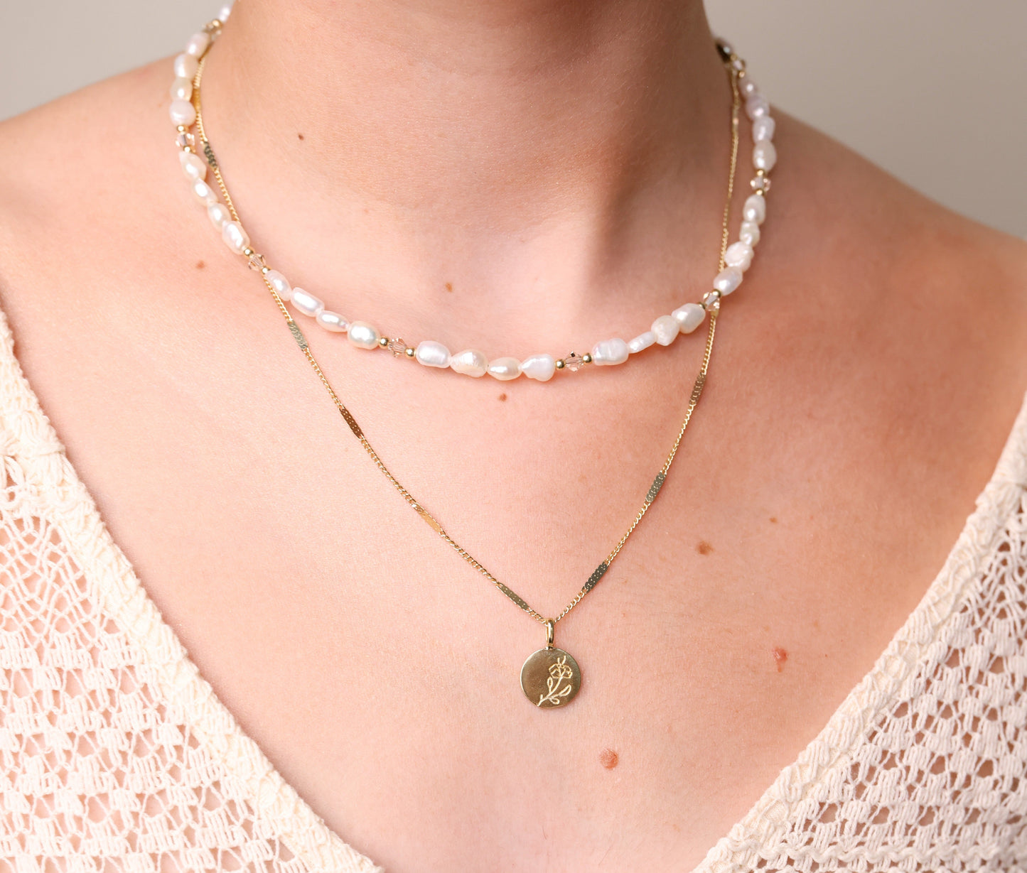 מבט תקריב על שרשראות ALAMOR BEIGE GOLD שכבות על צוואר דוגמנית, פנינים עם קריסטלים ותליון פרח זהב.