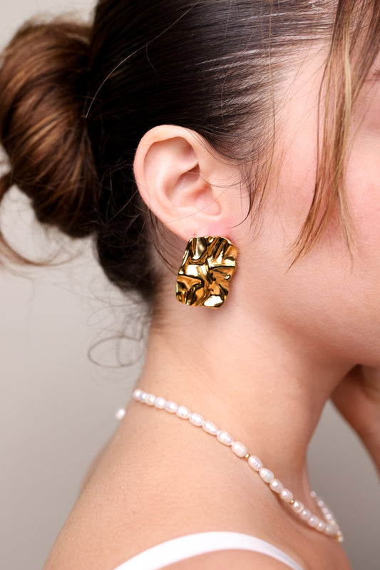 ART EARRINGS זהב מקומט על אוזן דוגמנית בפרופיל, שיער אסוף ושרשרת פנינים עדינה.