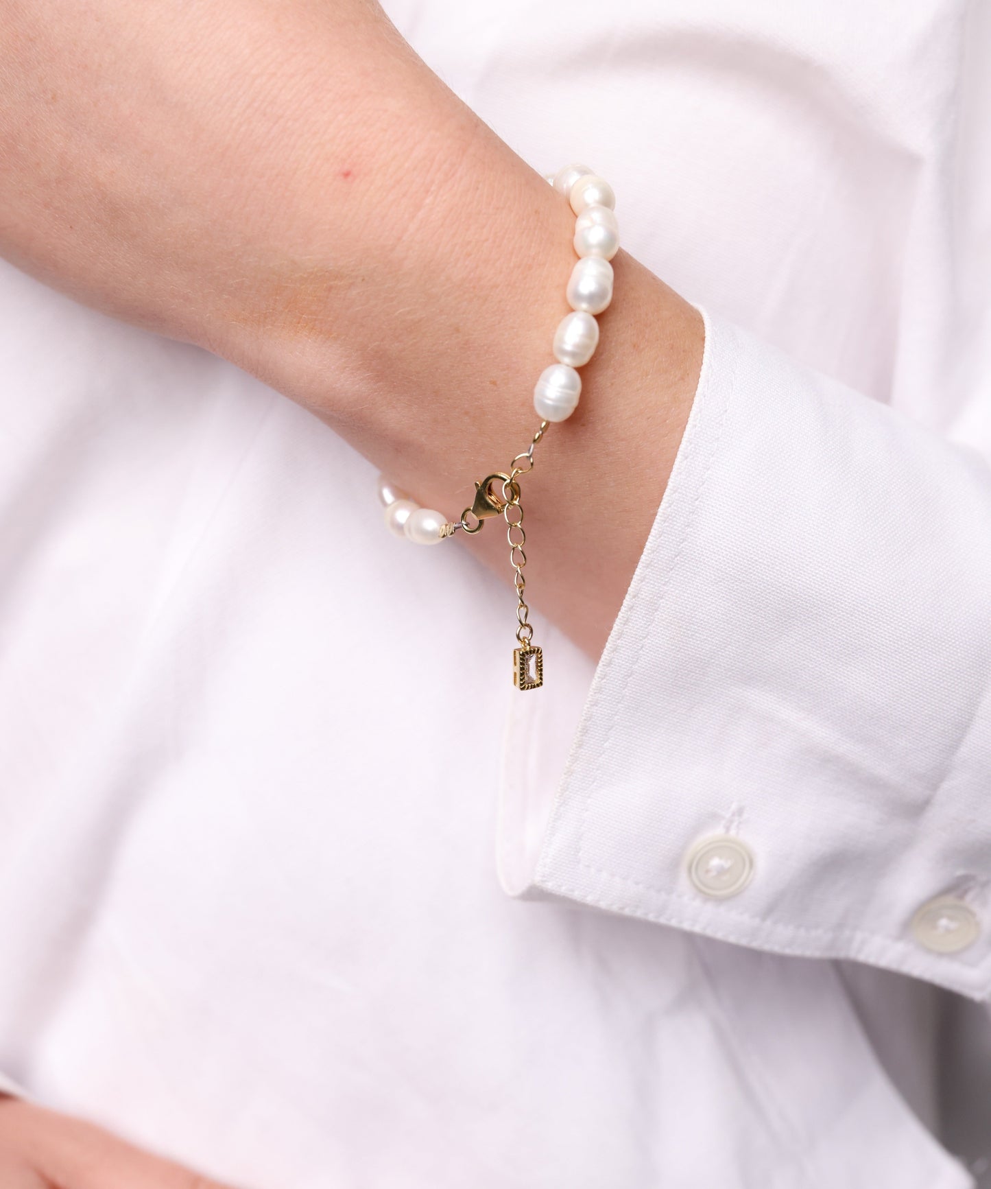 צמיד AMELIA פנינים לבנות אורגניות על פרק יד דוגמנית לובשת חולצה לבנה. תקריב על סוגר זהב עם תליון מלבני.