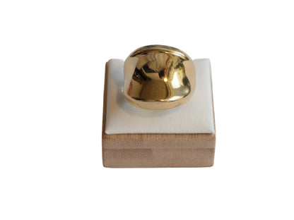 טבעת STAN RING זהובה, כיפה חלקה ומבריקה, מצולמת מקדימה על מעמד עץ עם כרית לבנה.