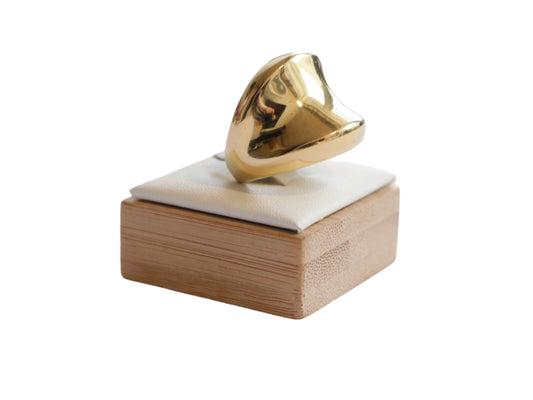 טבעת STAN RING מזהב מבריק בעיצוב גלי פיסולי, מוצגת על מעמד עץ עם כרית בהירה, מבט צד.