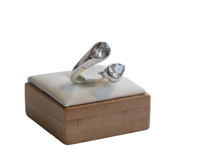 טבעת SHORE RING פתוחה בגוון כסף, משובצת בשתי אבנים נוצצות בגדלים שונים, על כרית תצוגה מעל עץ במבוק.