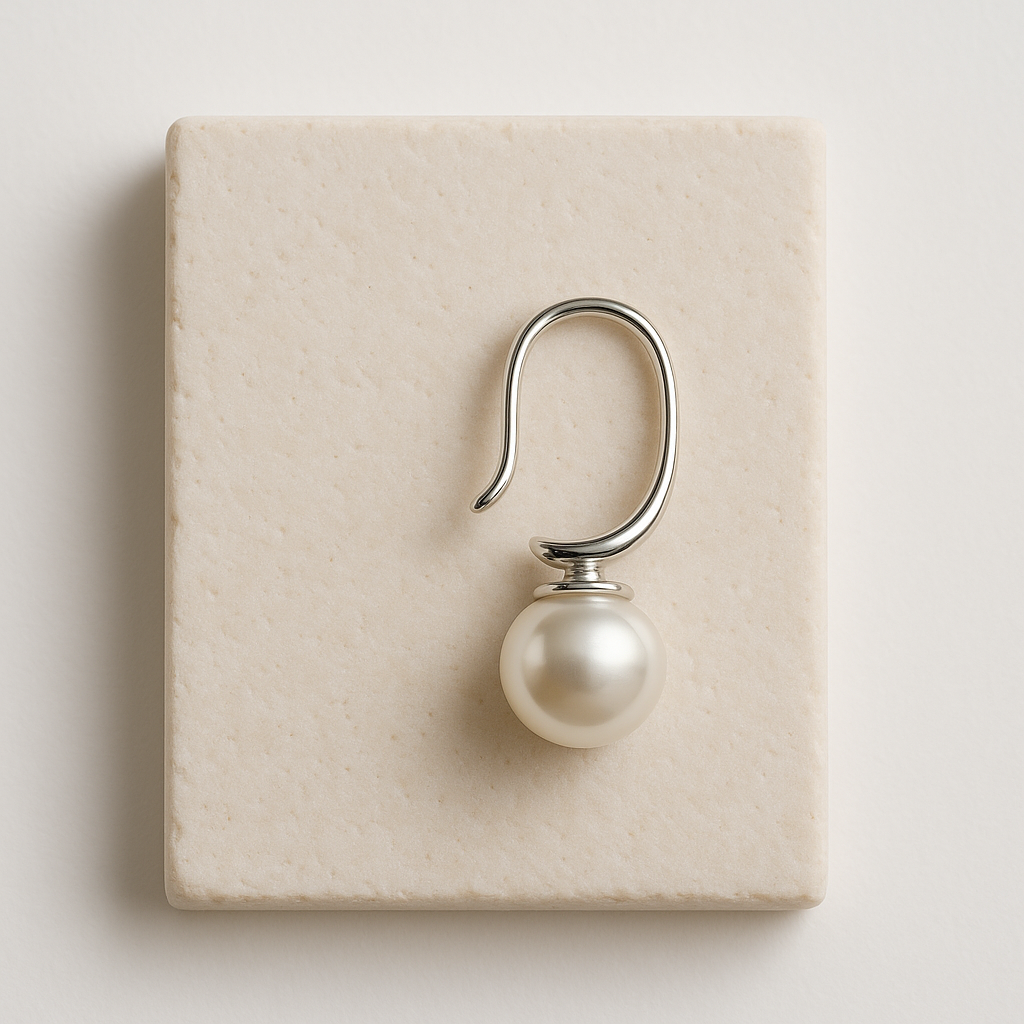 עגיל BERI EARRINGS יחיד, קשת כסף אלגנטית עם פנינה בוהקת, מונח על אריח אבן בהיר, תקריב מלמעלה.