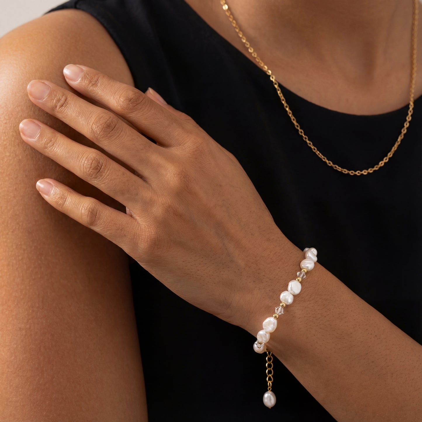 צמיד ALAMOR BEIGE GOLD אלגנטי, פניני בארוק וקריסטלים על פרק יד דוגמנית בפוזה טבעית, שרשרת זהב ברקע.